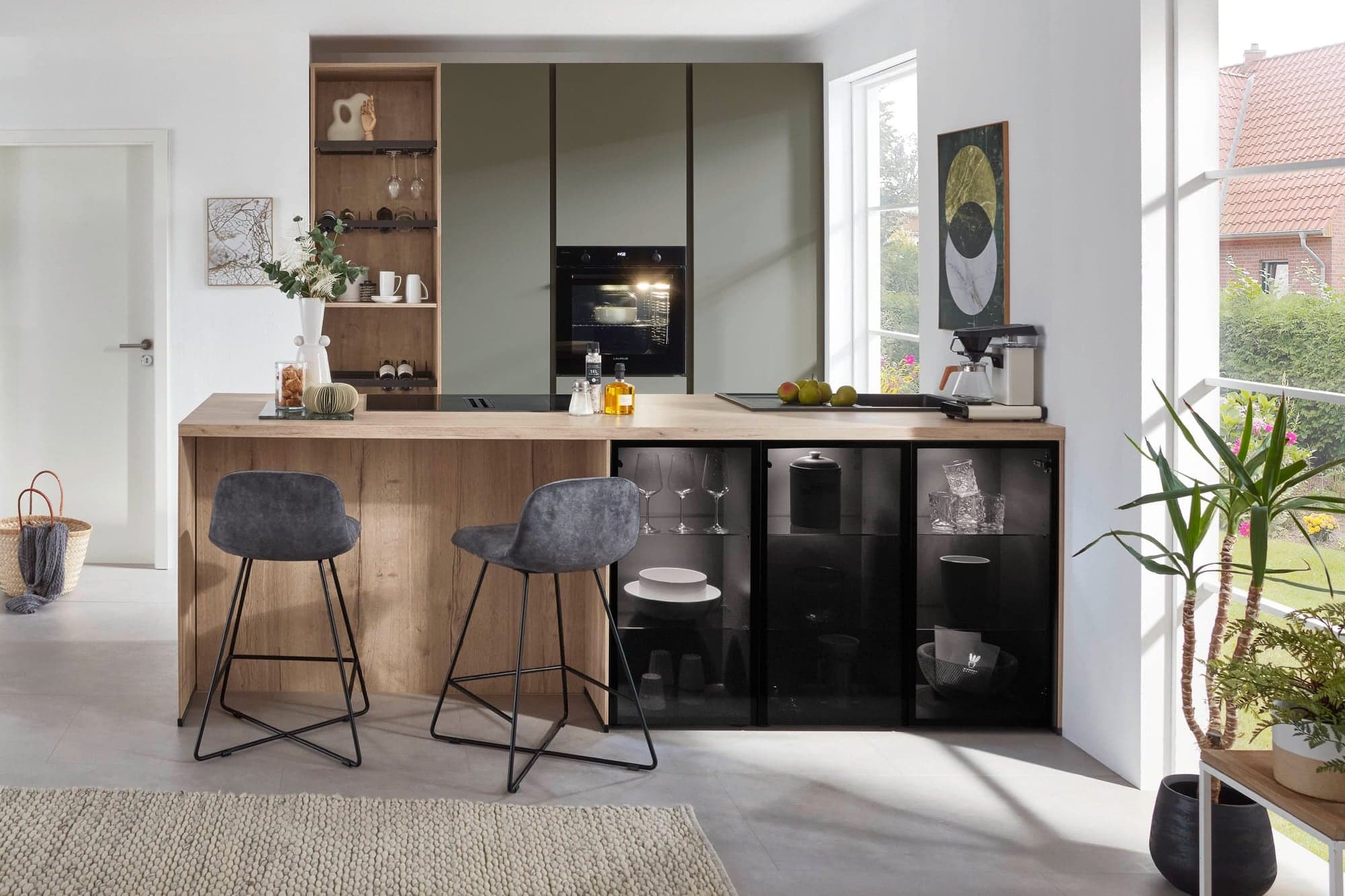 Offene Küche mit Theke in Eichenholz und zwei grauen Barhockern, olivgrünen Hochschränken mit beleuchtetem Backofen, schwarzen Glasvitrinen und einem hellen Fenster zum Garten