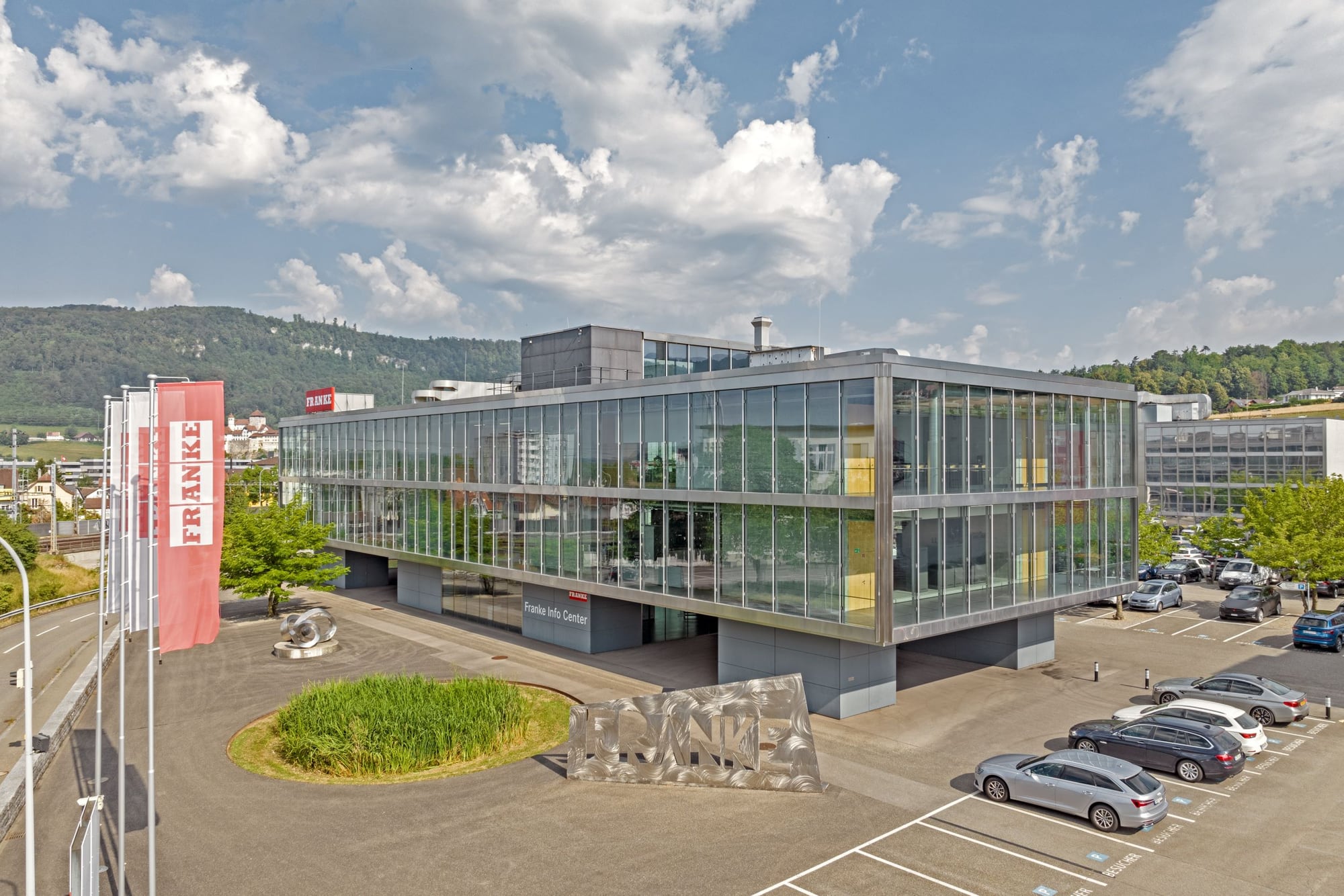Firmengebäude von FRANKE: ein Glasbau mit umlaufender Attika, davor rote Fahnen mit dem Markennamen und Parkplätze