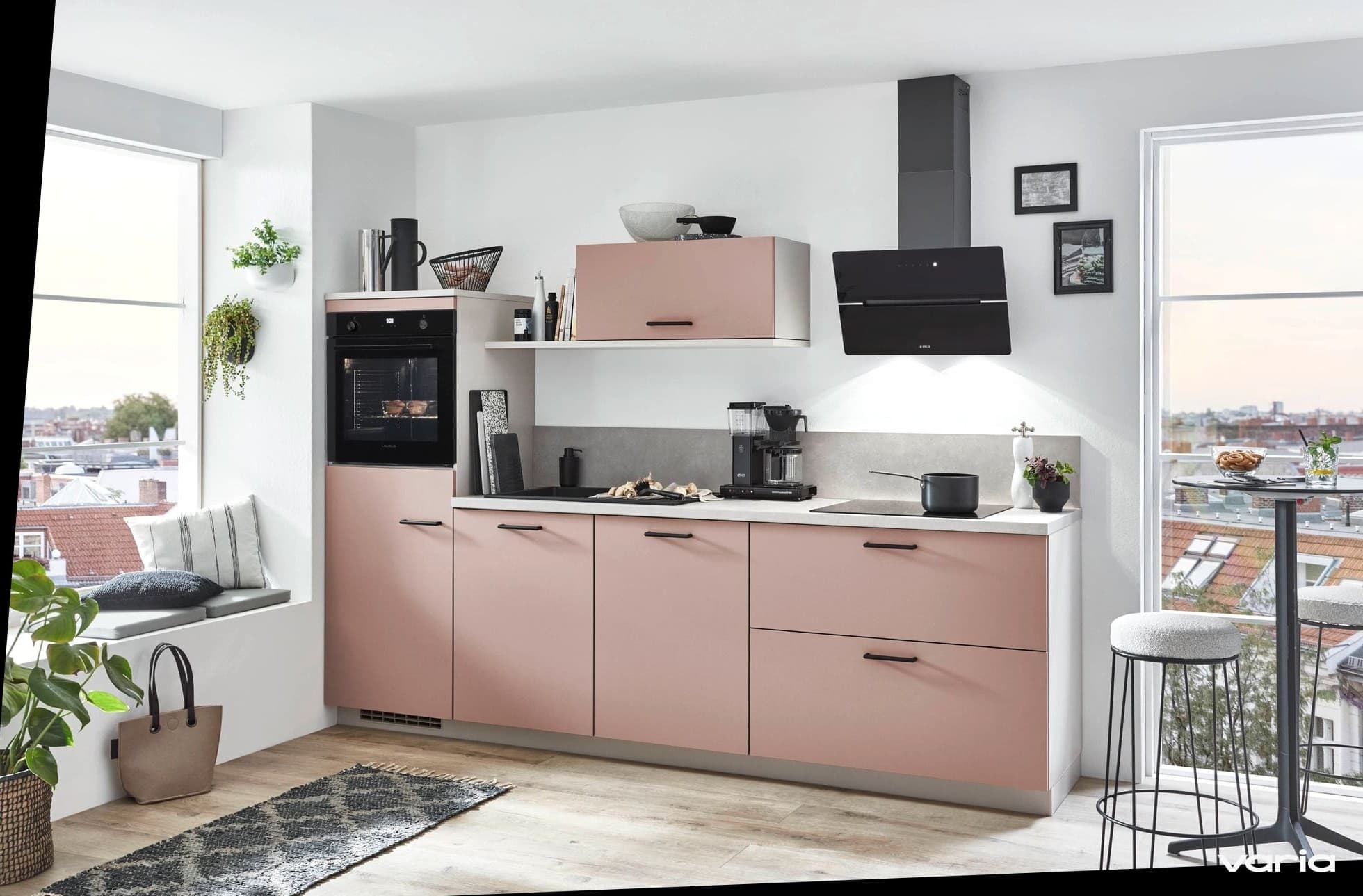 Helle Stadtküche mit altrosa Fronten und schwarzen Griffen: Backofen im Hochschrank, schwarze Schräghaube, Kaffeemaschine und Topf auf der hellen Arbeitsplatte, daneben eine Fensterbank mit Kissen und Pflanzen und Blick über die Dächer