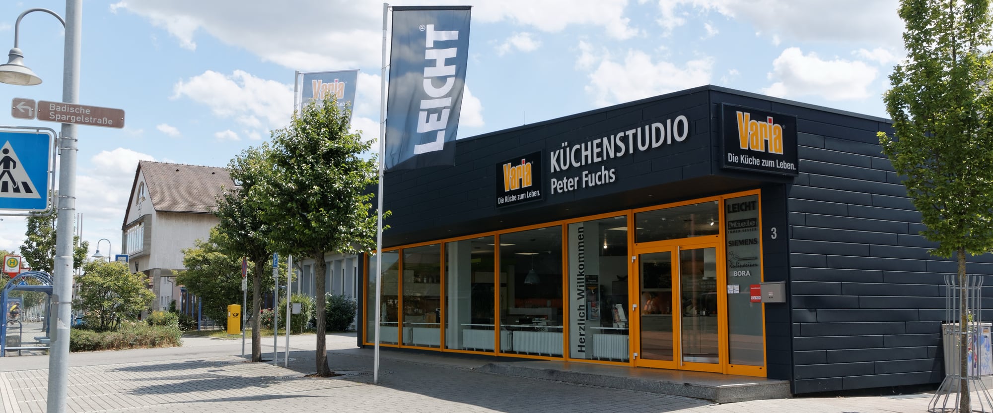 Küchenstudio Peter Fuchs in Karlsdorf von der Straße aus gesehen: dunkle Fassade mit Varia-Schriftzug, orange gerahmte Schaufenster und Eingang unter blauem Himmel