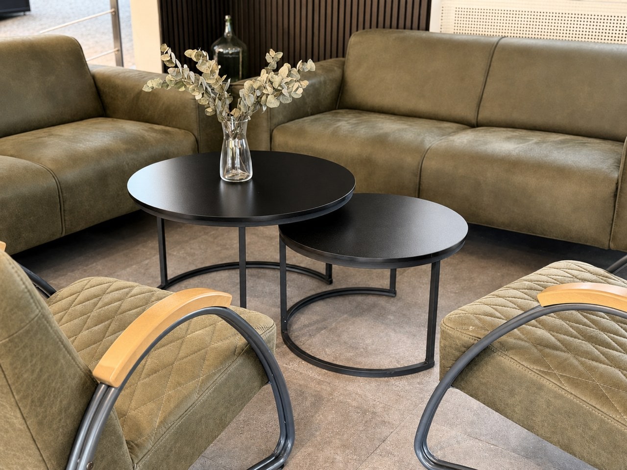 Sitzecke in der Ausstellung in Ludwigsburg: olivgrüne Ledersofas und zwei Sessel mit Holzarmlehnen um zwei runde schwarze Tische, darauf Eukalyptus in einer Glasvase