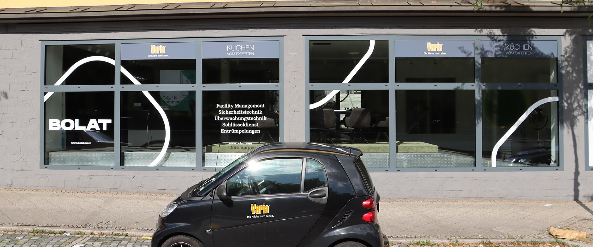 Das varia-Küchenstudio an der Stuttgarter Straße in Ludwigsburg von der Straße aus: lange graue Schaufensterfront mit geschwungenen weißen Linien auf den Scheiben, oben in den Fenstern die Tafeln mit der alten varia-Wortmarke und KÜCHEN VOM EXPERTEN, links die Beschriftung BOLAT, davor ein schwarzer Smart mit varia-Logo