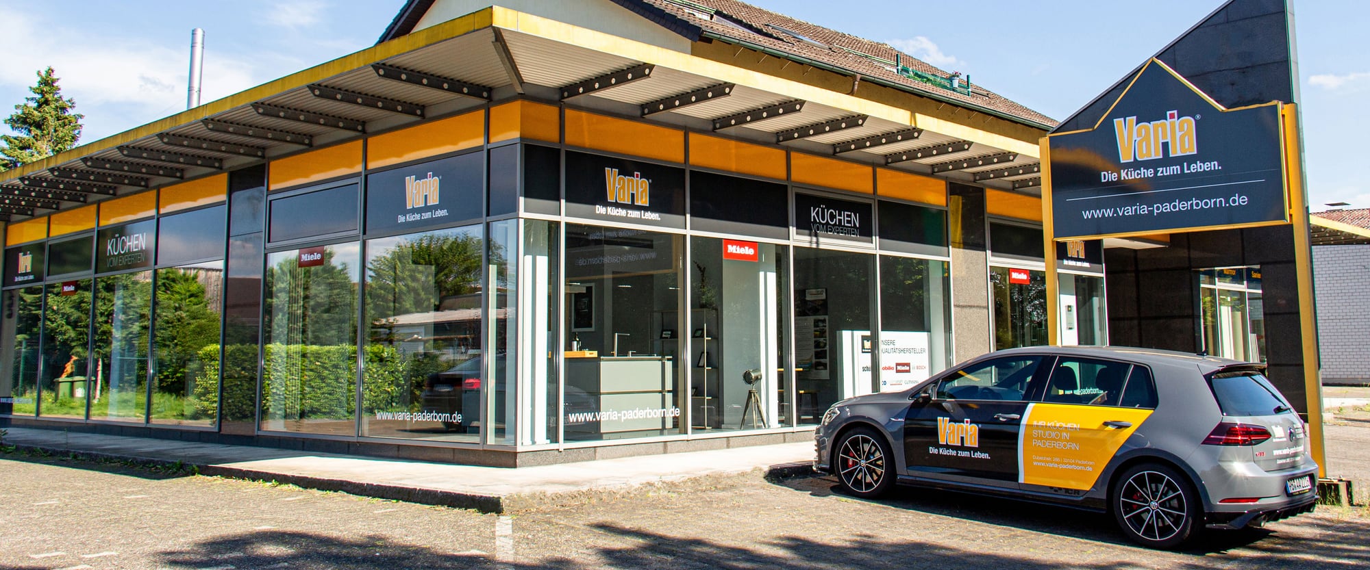 Das Küchenstudio in Paderborn-Schloß Neuhaus von der Straße aus: umlaufende Schaufensterfront unter dem gelben Vordach, rechts der Pylon, davor der Studiowagen