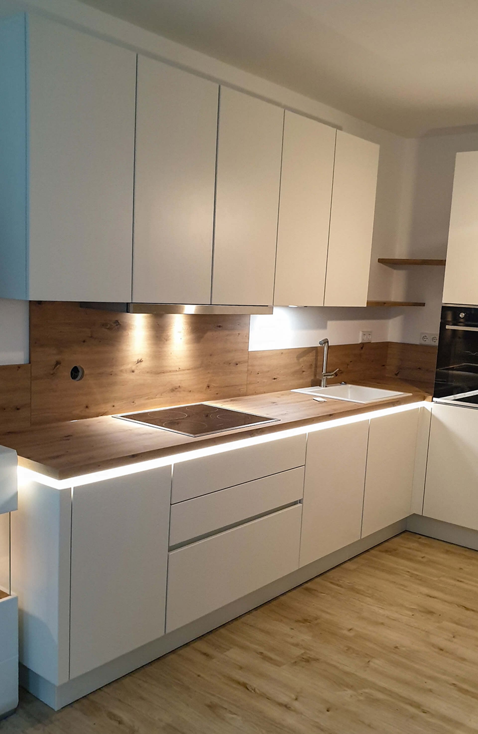 Fertig montierte grifflose Kundenküche in Weiß mit Eichen-Arbeitsplatte, Holzrückwand und Lichtband unter der Arbeitsplatte