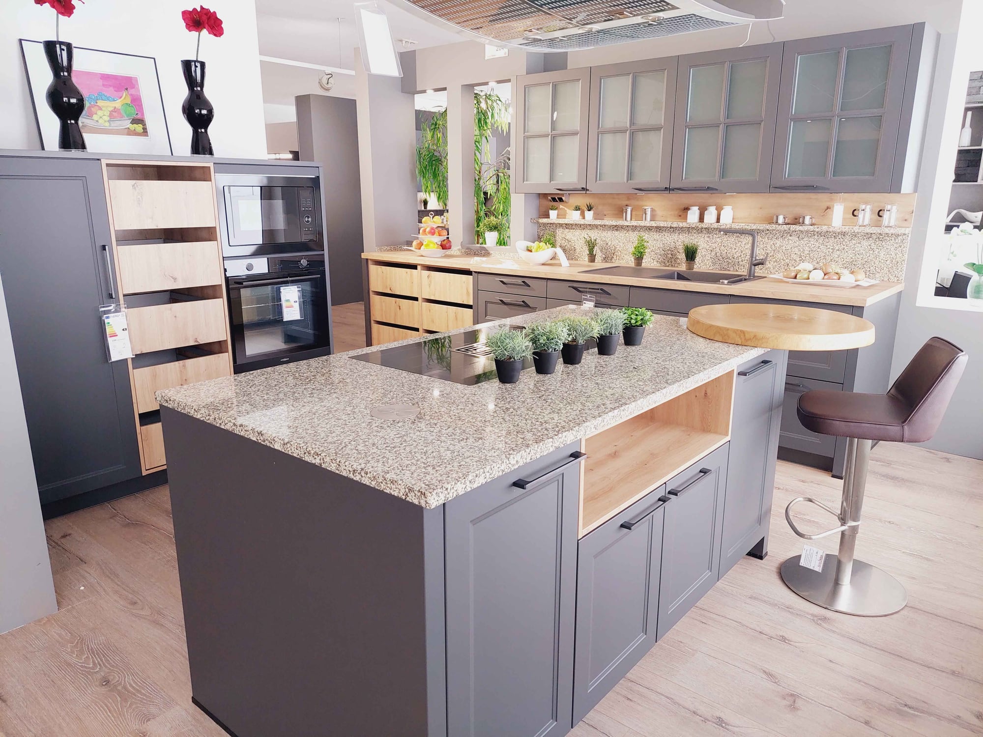 Ausstellungsküche in Weißdorf mit grauen Rahmenfronten, Insel mit gesprenkelter Platte in Granitoptik, Holzauszügen und Barhocker
