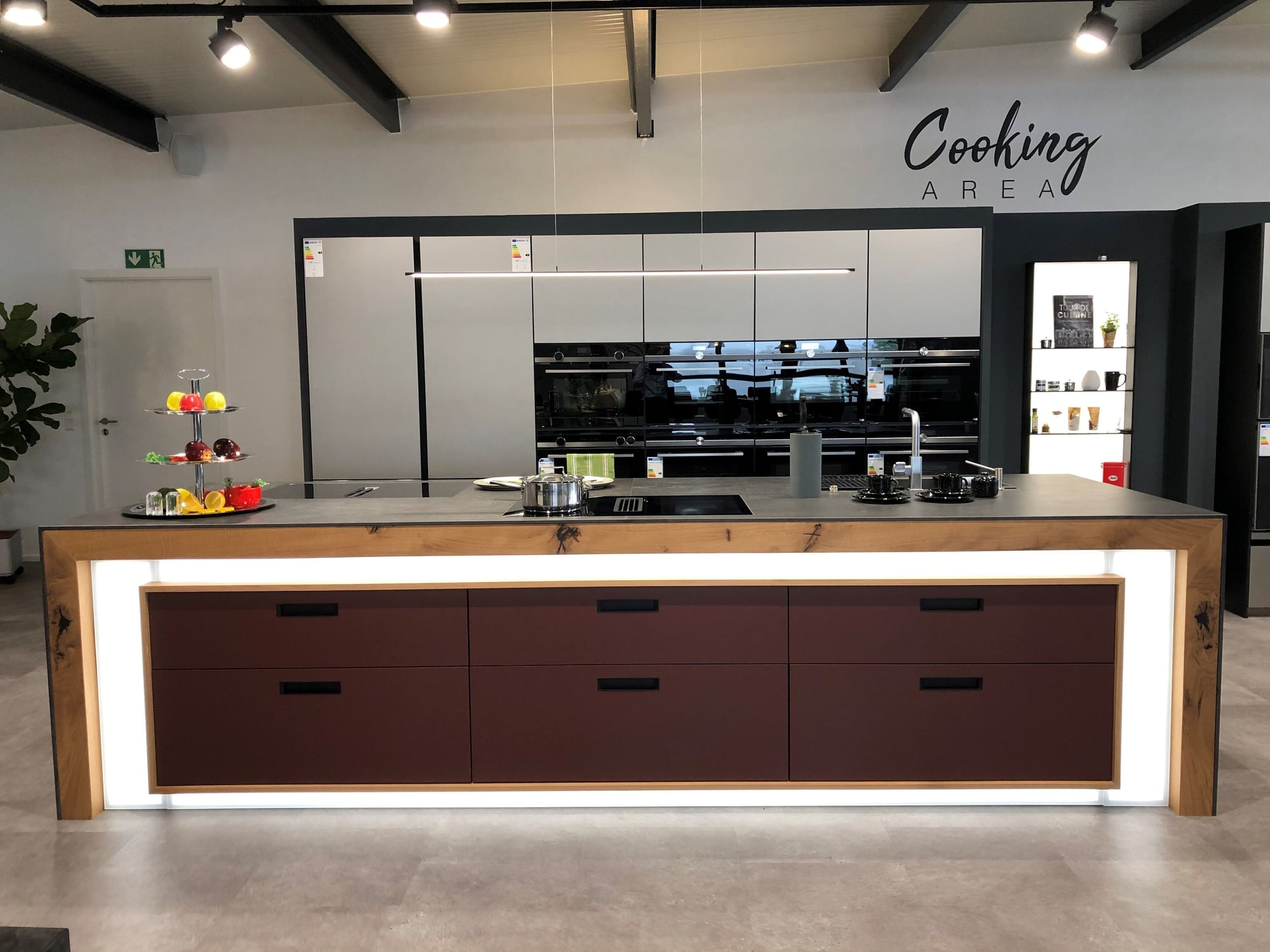 Cooking Area in Welzheim: Kochinsel mit dunkelroten Fronten in einem Eichenrahmen mit umlaufender Lichtkante, dahinter eine Zeile mit schwarzen Backöfen und der Schriftzug Cooking Area