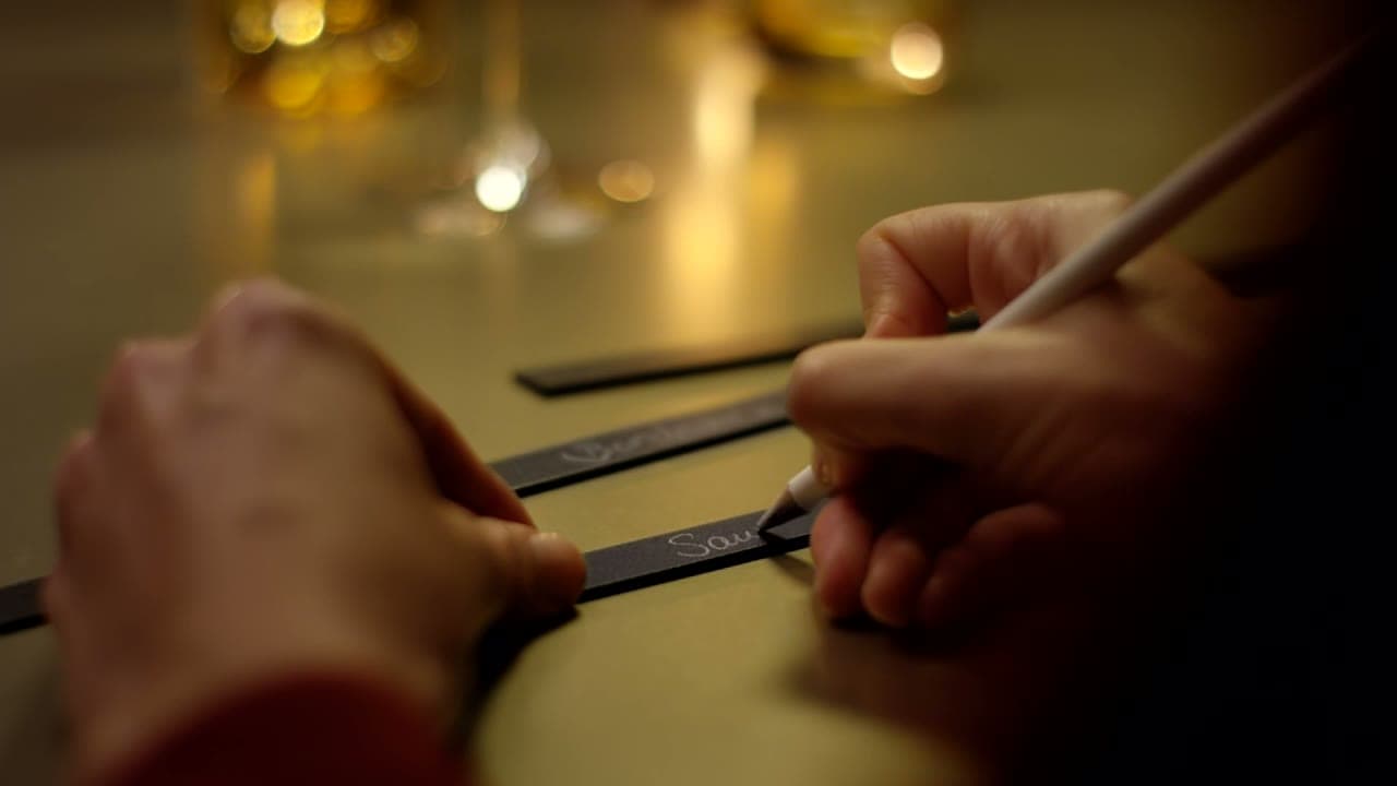 Eine Hand beschriftet eine schwarze Leiste mit Kreide und steckt sie an einen Holzrost mit Weißweinflaschen