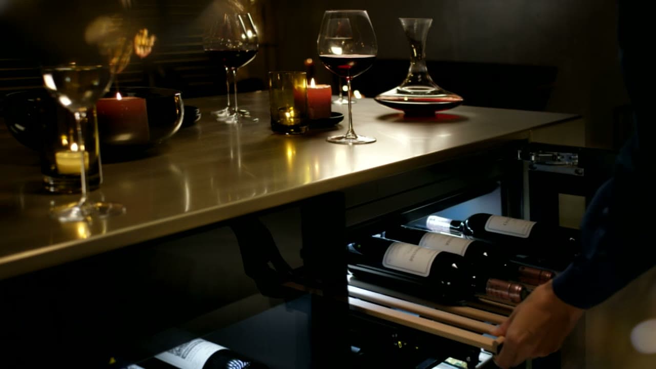 Dunkler Tresen mit Kerzen, Rotweingläsern und Karaffe, darunter ein geöffneter Weinschrank, aus dem eine Hand einen Holzrost mit Weinflaschen zieht